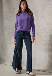 Pull violet côtelé avec un col bateau, associé à un jean droit bleu foncé. Le modèle tient un sac en denim et porte des baskets blanches.