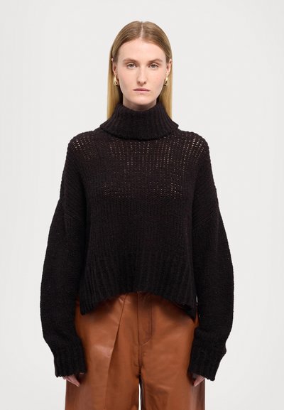 Schwarzer, verkürzter Pullover mit hohem, geripptem Rollkragen, lockerer Passform und strukturiertem Strickmuster, kombiniert mit braunen Lederhosen.