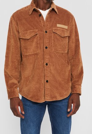 Chemise marron en velours côtelé à boutons avec deux poches poitrine, portée sur une chemise blanche et associée à un jean bleu.