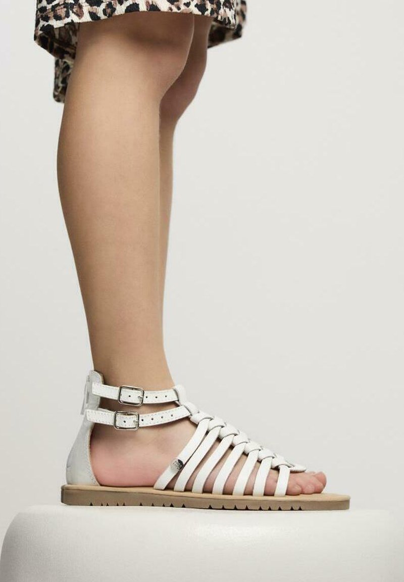 Sandali gladiatore in pelle bianca con molteplici cinturini, due fibbie alla caviglia, suola piatta color tan e design aperto sulla punta, esposti su un piedistallo bianco.