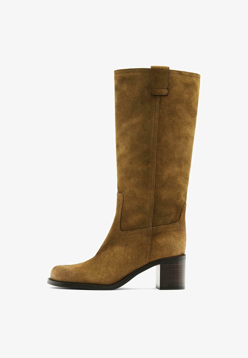 Botte en cuir suédine marron, hauteur genou, avec un bout carré et un talon bloc. Présente un panneau latéral et une languette en haut pour un enfilage facile.