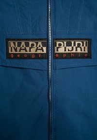 Chaqueta azul con dos parches rectangulares negros que presentan texto blanco y rojo, destacando los nombres de las marcas, centrados alrededor de la cremallera.