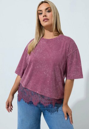 Donna con una maglietta viola larga e lavata con bordo in pizzo e jeans blu, in piedi contro uno sfondo chiaro e uniforme.