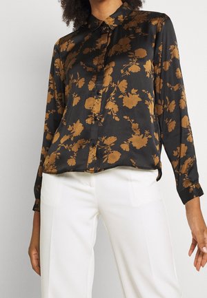 Mujer con camisa negra abotonada con patrón floral marrón y pantalones blancos de cintura alta, de pie frente a un fondo liso.