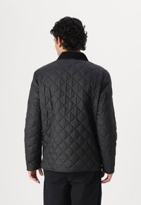 Casaco acolchoado preto com um padrão de diamantes, apresentando um colarinho suave, um ajuste descontraído e mangas longas. A vista traseira mostra detalhes de costura.