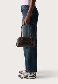 Väska med leopardmönster i glänsande material, med två handtag och dragkedjestängning, hålls av en person i blå jeans och mönstrade sneakers.