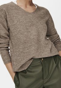Pull marron côtelé avec un col en V et des manches longues, associé à un pantalon en cuir vert, présentant une coupe décontractée et une texture douce.