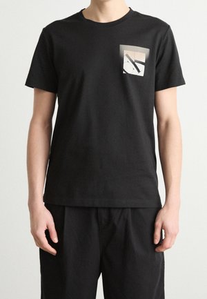 Personne portant une chemise noire à manches courtes avec un petit motif abstrait blanc et gris sur la partie supérieure gauche de la poitrine, associée à un pantalon noir.