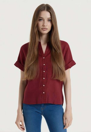 Giovane donna con lunghi capelli castani lisci che indossa una camicia rossa a maniche corte abbottonata e jeans blu, in piedi davanti a uno sfondo bianco.
