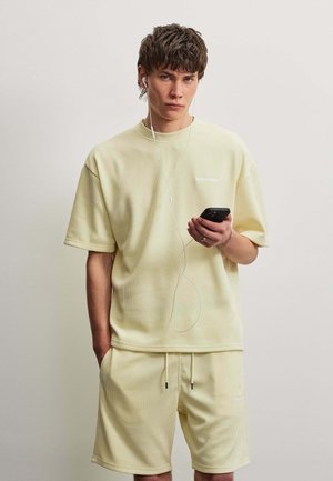 Junger Mann trägt hellgelbes, geripptes Kurzarmshirt und Shorts, hält ein Smartphone mit eingesteckten Ohrhörern vor einfarbigem Hintergrund.