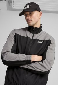 Chaqueta de entrenamiento PUMA con bloques de color negro y gris, cierre de cremallera, cuello alto y puños elásticos. La gorra presenta un logo en blanco.