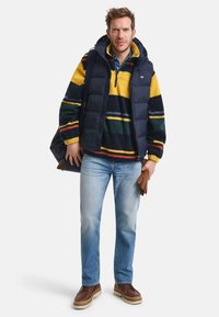 Dunkelblauer Puffer-Westen über einem mehrfarbigen gestreiften Fleece, getragen mit hellblauen Jeans und braunen Stiefeln. Hält braune Handschuhe und eine Tasche.