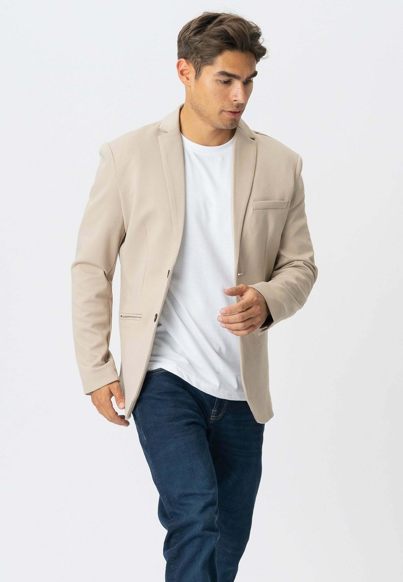 Beige blazer med en glat tekstur, enkelt brystlomme, to forlommer og en tætsiddende pasform, båret over en hvid t-shirt og blå jeans.