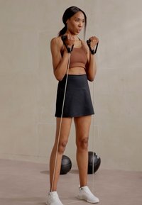 Modstandsbånd holdt af en person i en brun sportstop og sorte nederdele. Outfitet står i kontrast til de hvide sneakers og den lyse baggrund.