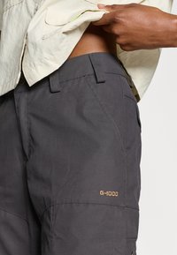 Grå G-1000 byxor har ett hållbart tyg, rak skärning, synliga fickor och guldbroderi. Designen inkluderar bälteshällor och en mjuk textur.