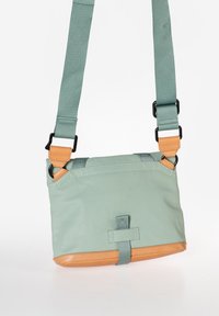 Borsa verde menta con una texture liscia, fondo in pelle marrone chiaro, tracolla regolabile e accessori neri. Presenta un dettaglio con fibbia frontale e un design minimalista.
