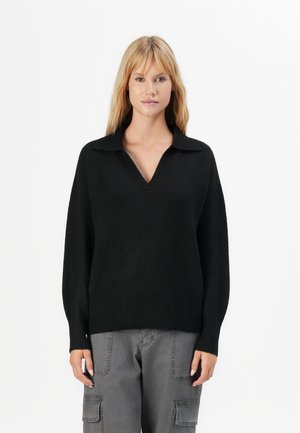 Vrouw in een zwart langmouw V-hals gebreid sweater en grijze cargobroek, staande tegen een eenvoudige witte achtergrond.