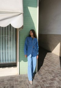 Jeansjacke und weit geschnittene Jeans in Hellblau, mit lockerer Passform. Glatte Textur mit kontrastierenden Nähten, vor einer grünen Fliesenwand.