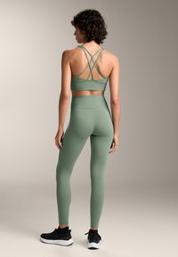 Groene sportbeha met kruislings straps en bijpassende high-waisted leggings, gemaakt van een gladde stof. Zwarte sportschoenen maken de outfit compleet.