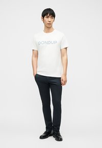 Homme debout portant un t-shirt blanc "DONDUP", un pantalon slim foncé et des chaussures habillées noires, avec une main dans la poche.