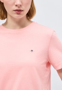 Γυναίκα που φοράει ανοιχτό ροζ κοντομάνικη μπλούζα με ένα μικρό κόκκινο, άσπρο και μπλε σήμα σε σχήμα ασπίδας στο πάνω αριστερό μέρος του στήθους.