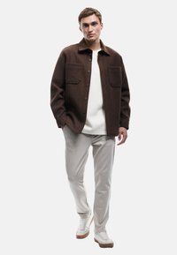 Mann in einer braunen knöpfbaren Jacke über einem weißen Hemd, hellgrauen Hosen und weißen Sneakern, der mit teilweise in den Taschen versenkten Händen steht.