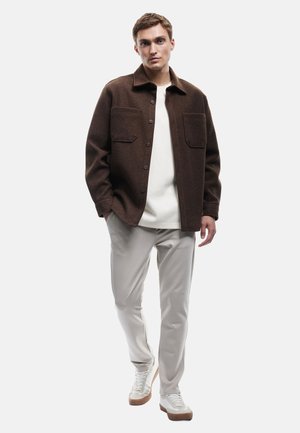 Mann in einer braunen knöpfbaren Jacke über einem weißen Hemd, hellgrauen Hosen und weißen Sneakern, der mit teilweise in den Taschen versenkten Händen steht.