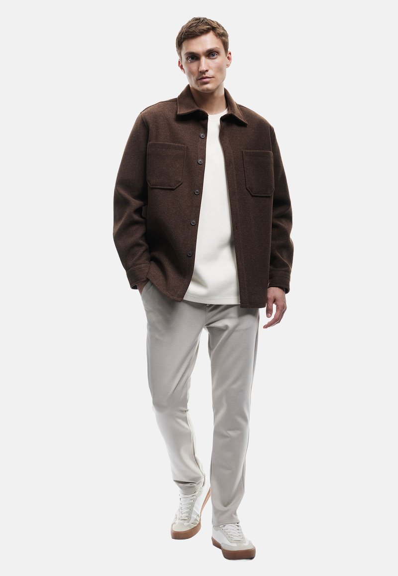 Mann in einer braunen knöpfbaren Jacke über einem weißen Hemd, hellgrauen Hosen und weißen Sneakern, der mit teilweise in den Taschen versenkten Händen steht.