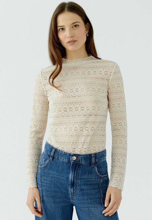 Junge Frau mit langen braunen Haaren trägt ein cremefarbenes Spitzen-Longsleeve und blaue Jeans, steht vor einem schlichten Hintergrund.
