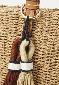 Borsa in rafia intrecciata con tracolla in pelle marrone e dettagli in oro. Accenti con frange in marrone, crema e nero aggiungono texture e dettaglio.
