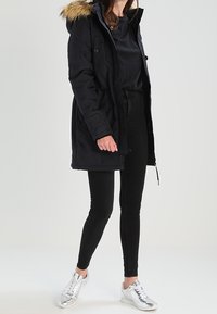 Kvinna klädd i svart parkas med pälskrage, svarta byxor, svart topp och silverfärgade sneakers, stående mot en neutralt bakgrund.