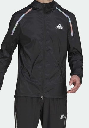 Mann trägt schwarze Adidas-Sportjacke und Hose mit reflektierenden Streifen an den Schultern sowie Adidas-Logos auf Brust und Hose.