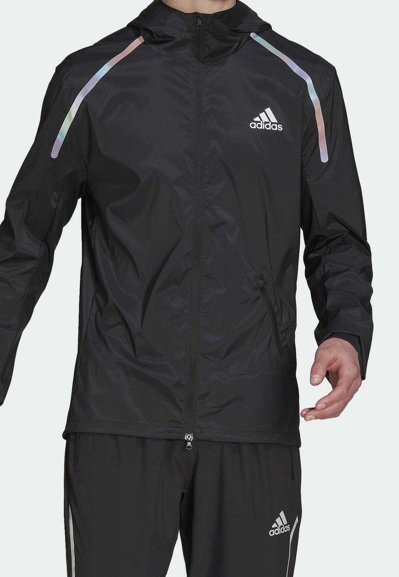 Homme portant une veste de sport noire Adidas et un pantalon assorti avec des bandes réfléchissantes sur les épaules et des logos Adidas sur la poitrine et le pantalon.