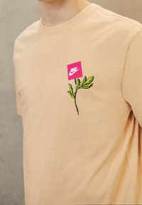 Beżowa bawełniana koszulka z różowym kwadratem z logo Nike oraz ilustracją zielonej rośliny na lewej piersi.