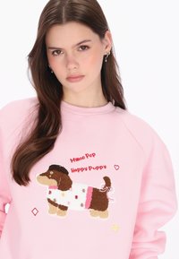 Rózsaszín pulóver egy texturált, rajzfilmfigurás kutya applikációval. A design élénk akcentusokat és játékos szöveget tartalmaz, amely így szól: "Myro Pop Boldog Kiskutya."