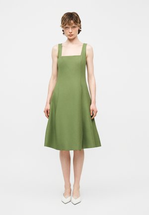 Žena stojaca čelom, oblečená v bezrukávových midi šatách olivovo zelenej farby so štvorcovým výstrihom a bielych topánkach na podpätku s ostrou špičkou na bielom pozadí.