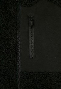 Schwarze Fleecejacke mit strukturierter Oberfläche, glatten Stoffakzenten und einer mit Reißverschluss versehenen Brusttasche mit schwarzem Zubehör.