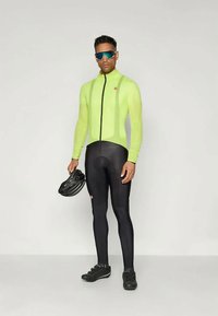 Cyklistický outfit pozostávajúci z priliehavého, jasne žltého dlhého cyklistického dresu a čiernych legín, s helmou držanou v jednej ruke a slnečnými okuliarmi.