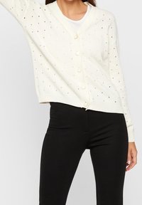 Kvinna klädd i en vit knäppbar stickad cardigan med hålmönster över en vit topp, i kombination med högmidjade svarta tajta byxor.