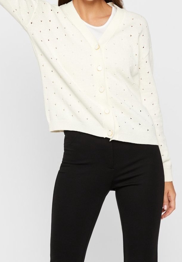 Kvinna klädd i en vit knäppbar stickad cardigan med hålmönster över en vit topp, i kombination med högmidjade svarta tajta byxor.