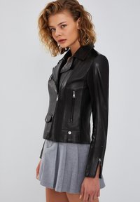 Giacca biker in pelle nera con un design aderente, tasche con zip e hardware in argento. Caratterizzata da un colletto a scollatura e una texture liscia.