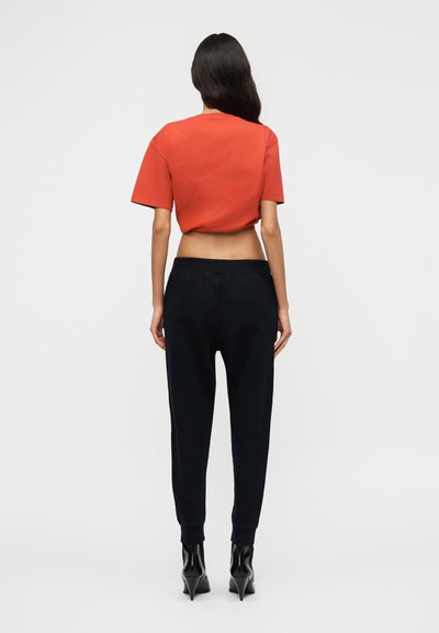 Γυναίκα με μακριά σκούρα μαλλιά, ντυμένη με κοντό κόκκινο μπλουζάκι, μαύρο φαρδύ παντελόνι με ψηλή μέση και μαύρα μποτάκια με τακούνι, στέκεται γυρισμένη προς τα πίσω.