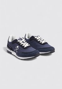 Un par de zapatillas azul marino y negras con cordones blancos y un logo en el lateral, exhibidas sobre un fondo gris claro liso.