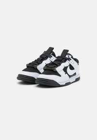 Scarpe da ginnastica bianche e nere con una punta nera testurizzata, lacci neri e un prominente logo swoosh nero sul lato. Suola in gomma.