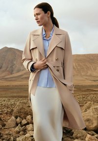 Beżowy płaszcz typu trench z podwójnym zapięciem, szerokimi klapami i paskiem do zawiązania. Nosi się go na jasnoniebieskiej bluzce i białej spódnicy; gładkie faktury.