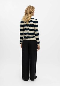 Gestreifter Pullover in Creme und Marineblau mit gerippter Textur und Zickzackmuster. Kombiniert mit lockeren schwarzen Hosen und flachen Schuhen.