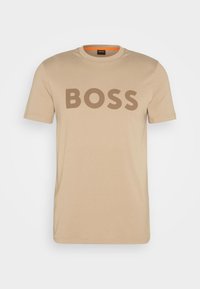 Beige bomulls-t-shirt med korta ärmar som har "BOSS" i tjocka bruna bokstäver över bröstet. Rund halsringning med en orange detalj inuti.
