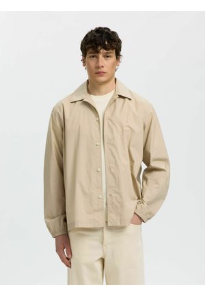 Jeune homme aux cheveux bouclés et foncés portant une veste beige à boutons par-dessus une chemise claire, debout avec une main dans la poche devant un fond uni.