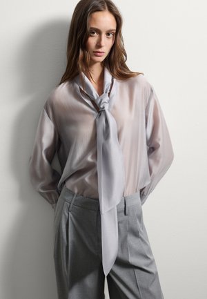 Lichtgrijze, doorschijnende blouse met een strik aan de voorkant en lange mouwen, gecombineerd met hooggeposeerde, op maat gemaakte grijze broeken met zijzakken.