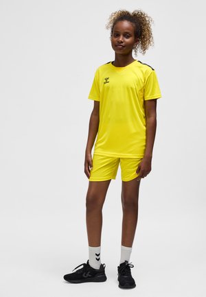 Gul sportstøj bestående af en kortærmet trøje og shorts. Glat, letvægtsstof med sorte accenter på skuldrene. Båret med sorte sko og grå sokker.